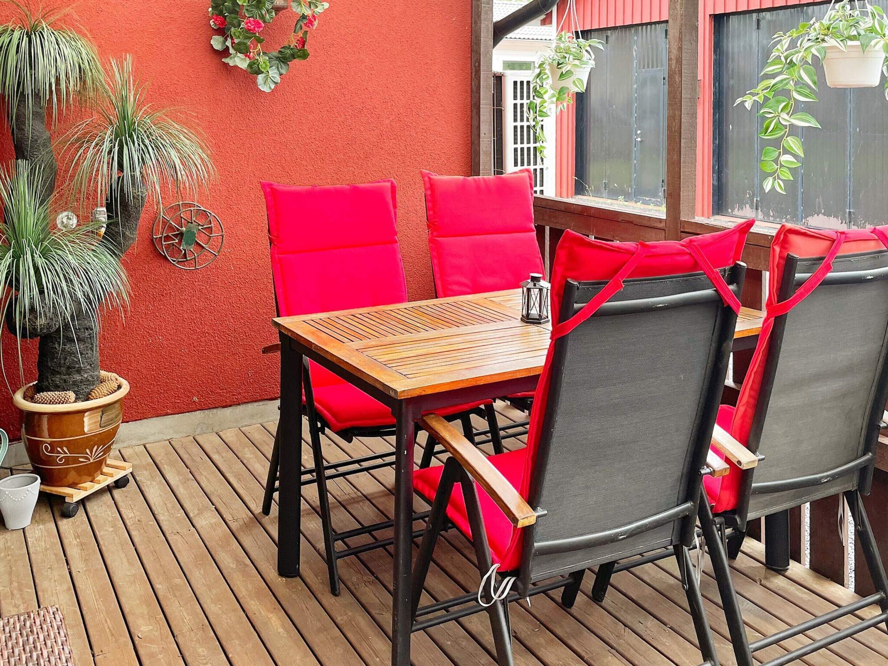 An outdoor patio with a wooden table and red cushioned chairs, surrounded by potted plants and a red wall.