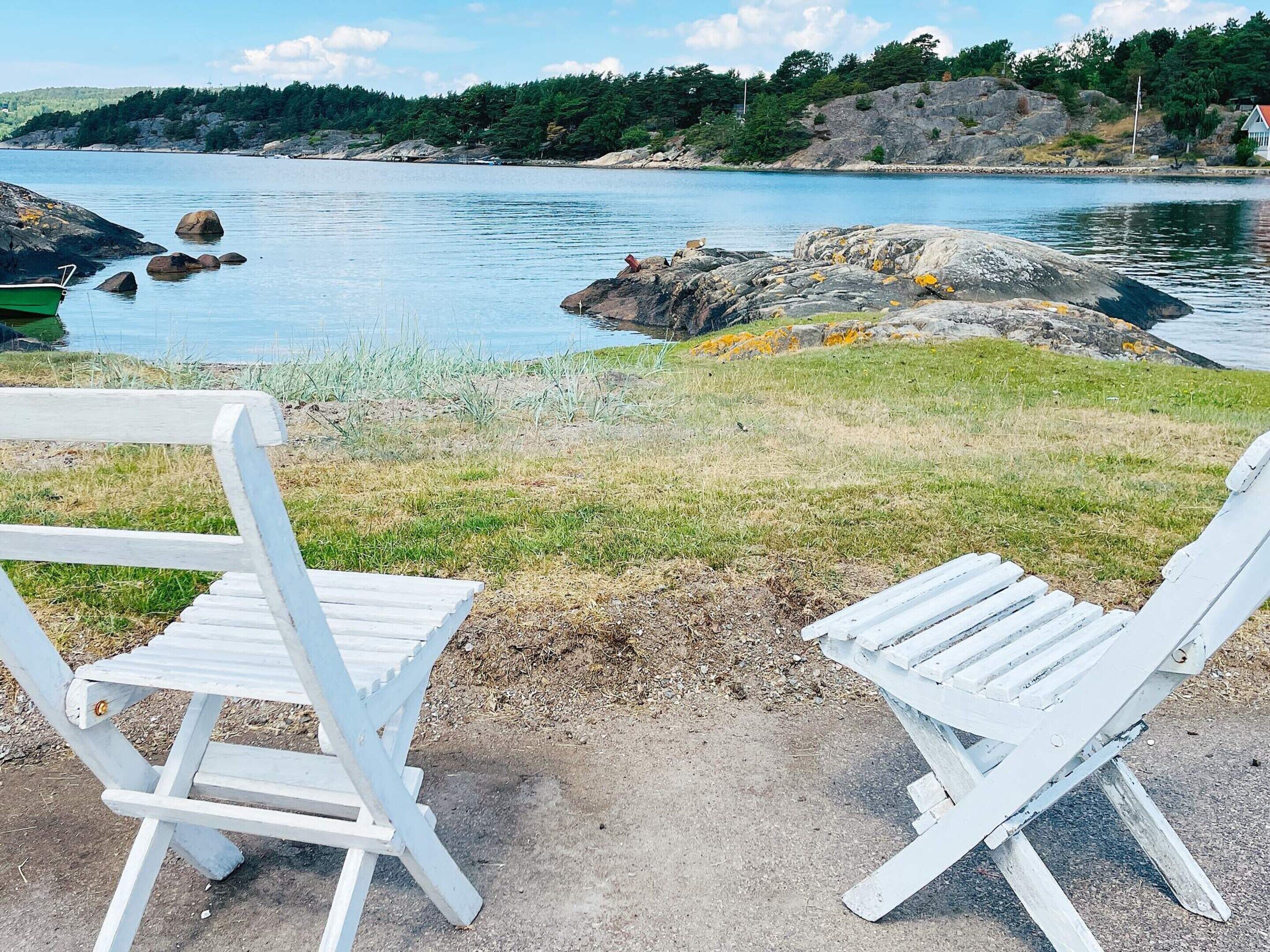 Two white Adirondack chairs facing a lake with rocky shores and a forested hill in the background.