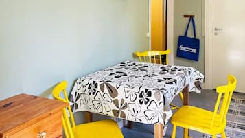 A room with a table covered in a patterned tablecloth, two yellow chairs, a wooden chest, and a blue bag hanging on the wall.