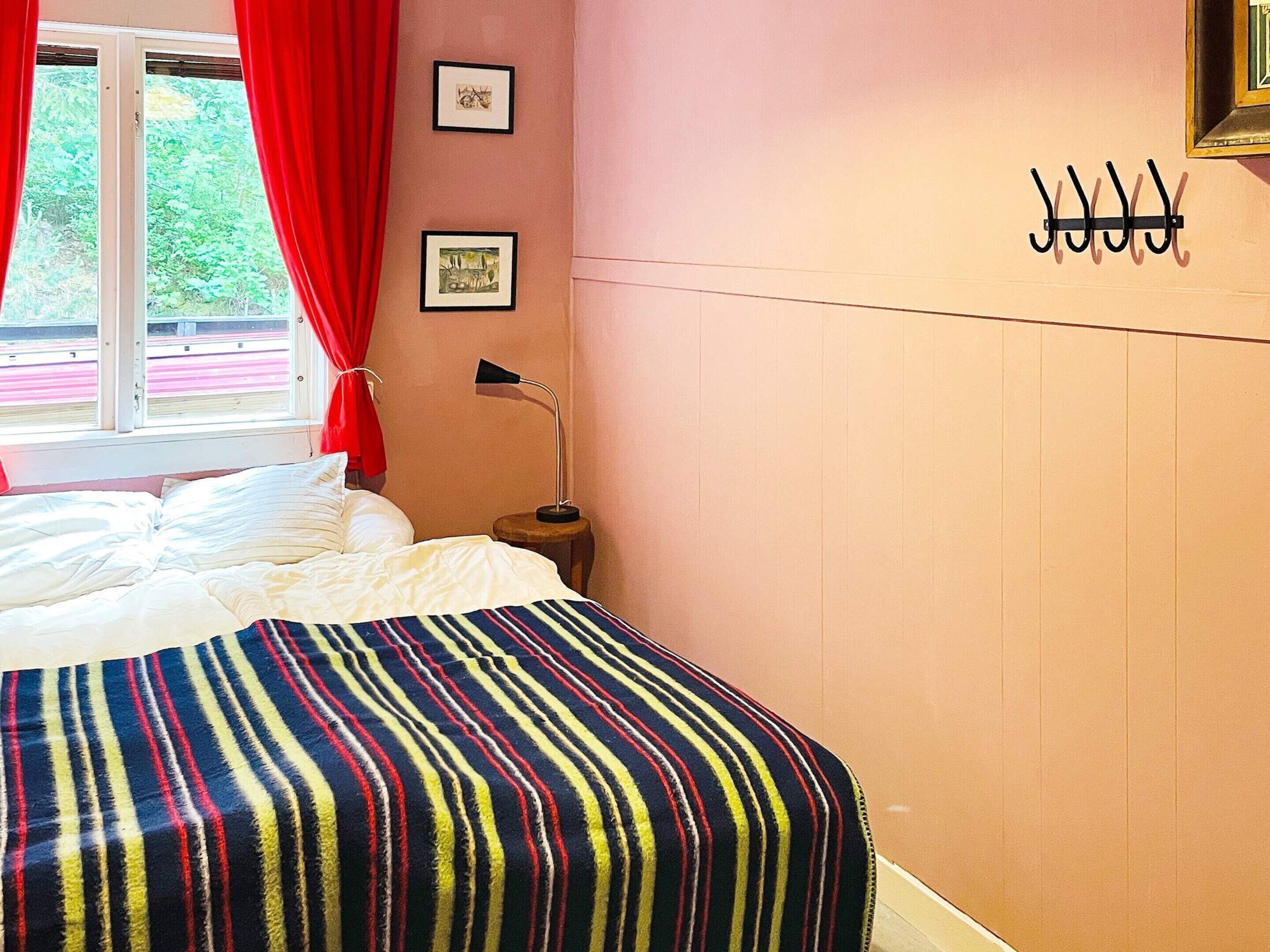 Quarto com cama, janela com cortinas, luminária de cabeceira e cabideiro fixado na parede.