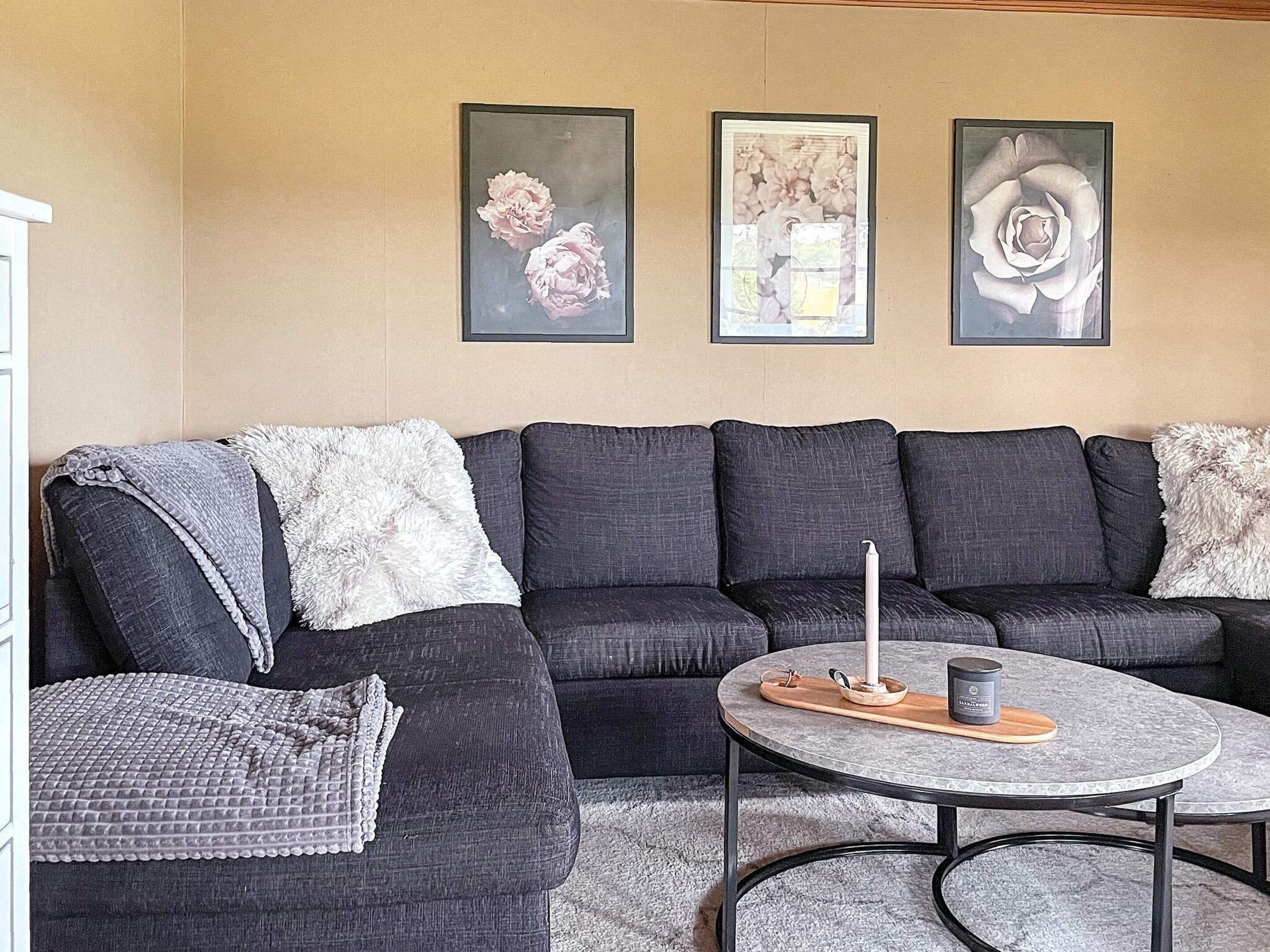 A living room with a dark grey sectional sofa, a round coffee table with a candle, and three framed floral artworks on the wall.