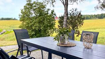 An outdoor dining area with a table and chairs, surrounded by greenery and a view of a field.