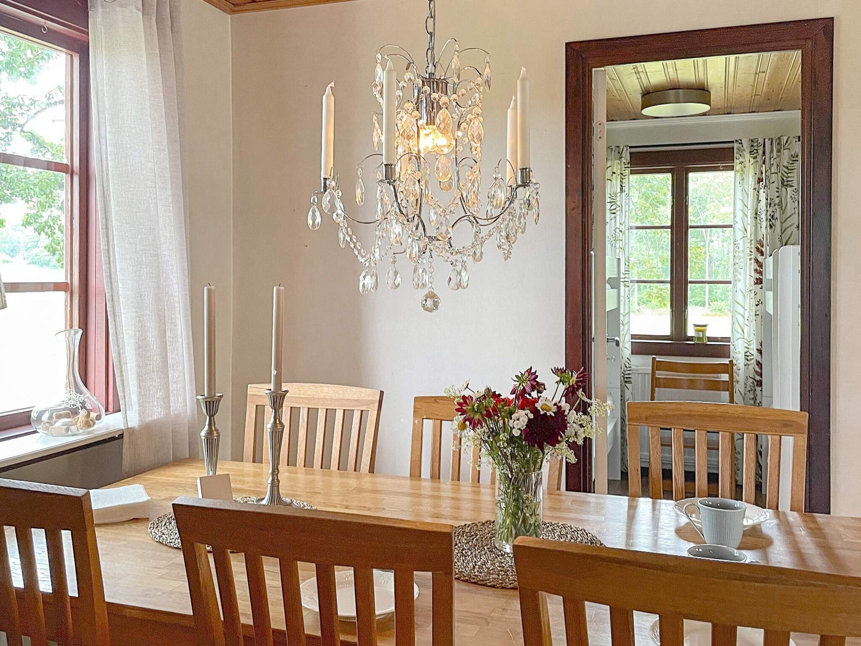 A dining room with a chandelier, wooden chairs, a table with a vase of flowers, and a window with curtains.