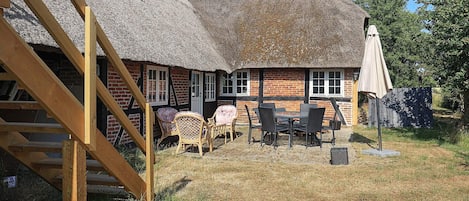 A thatched-roof house with a wooden staircase, a dining area with chairs and a table, and a covered outdoor seating area.