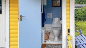 A small, single-room structure with a blue interior, a white toilet, a framed picture on the wall, and a yellow umbrella outside. - 4 Person Holiday Home in Kolmarden (Kolmården)
