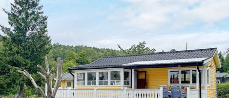 Keltainen mökki, jossa on valkoinen kuisti ja katettu terassi, ja sen ympärillä valkoinen puinen aita. Mökki sijaitsee ruohikkoisella niityllä, jonka taustalla näkyy puita.