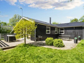 A building with a black exterior, a sloped roof, and a chimney. There is a paved area with a bench and a table, surrounded by greenery. - 6 Person Holiday Home in Hemmet (Hemmet)