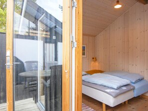 A wooden cabin with a glass door leading to an outdoor area with a table and chairs. Inside, there is a bed with two pillows and a small bedside table with a lamp.