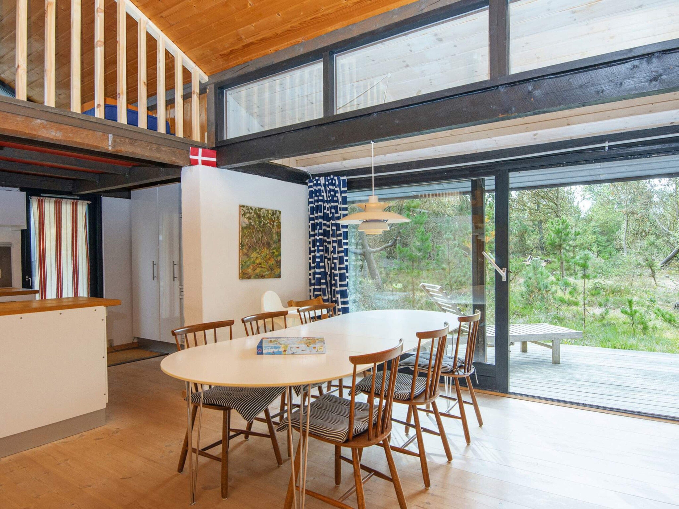 Uma sala de jantar com mesa redonda e seis cadeiras, um quadro na parede e vista para o exterior através de grandes janelas.