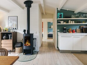 A cozy living room with a wood-burning stove, a dining area with a table and chairs, and a kitchen with shelves of glassware and kitchenware. - 4 Star Holiday Home in Stege (Stege)