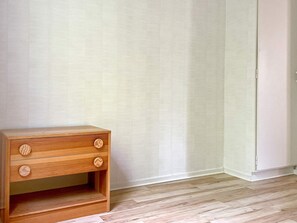 Une table de chevet en bois, dotée de tiroirs et d’une étagère, placée contre un mur recouvert d’un papier peint à motifs simples.