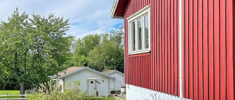 Un bâtiment rouge avec une fenêtre blanche et une porte blanche, entouré de verdure et bordé d’un chemin de gravier.
