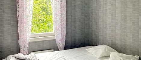 A bedroom with a bed, white bedding, a window with curtains, and a view of greenery outside.