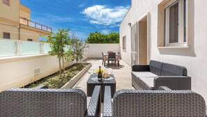 Eine Terrasse im Freien mit Korbmöbeln, einem Tisch mit Flasche und Gläsern und Blick auf ein Gebäude im Hintergrund.