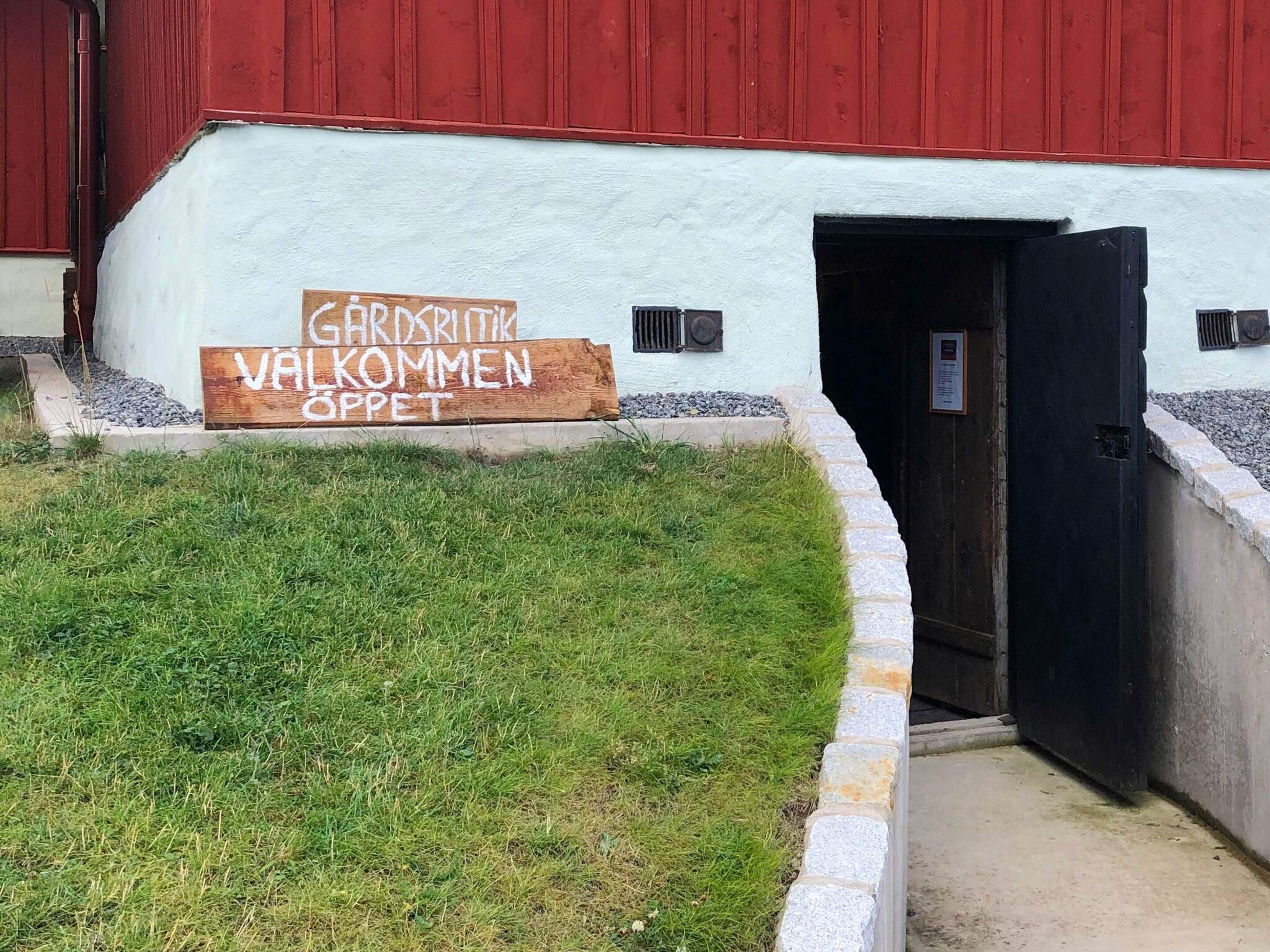 A building with a white exterior and a red door. There is a wooden sign in front of the building that reads 'GARDENBRIEFT WAKOMMEN SPPET'.