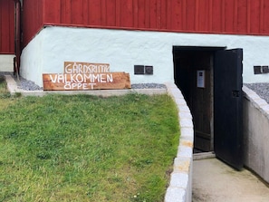 Un bâtiment à l’extérieur blanc et à la porte rouge. Devant le bâtiment, on peut voir une enseigne en bois sur laquelle on peut lire : « GARDENBRIEFT WAKOMMEN SPPET ».
