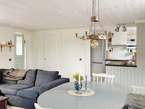 A modern living room with a grey sofa, a round coffee table with a vase, and a kitchen area with white cabinets and a stainless steel refrigerator. - 4 Star Holiday Home in Mellosa (Flen)