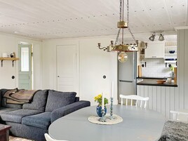 Una sala moderna con un sofá gris, una mesa redonda con un florero, y una zona de cocina con armarios blancos y un refrigerador de acero inoxidable.