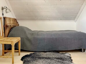 Une chambre à coucher aménagée dans un grenier, avec un plafond en pente, un cadre de lit en bois, un matelas gris, une petite table en bois et un tapis gris.