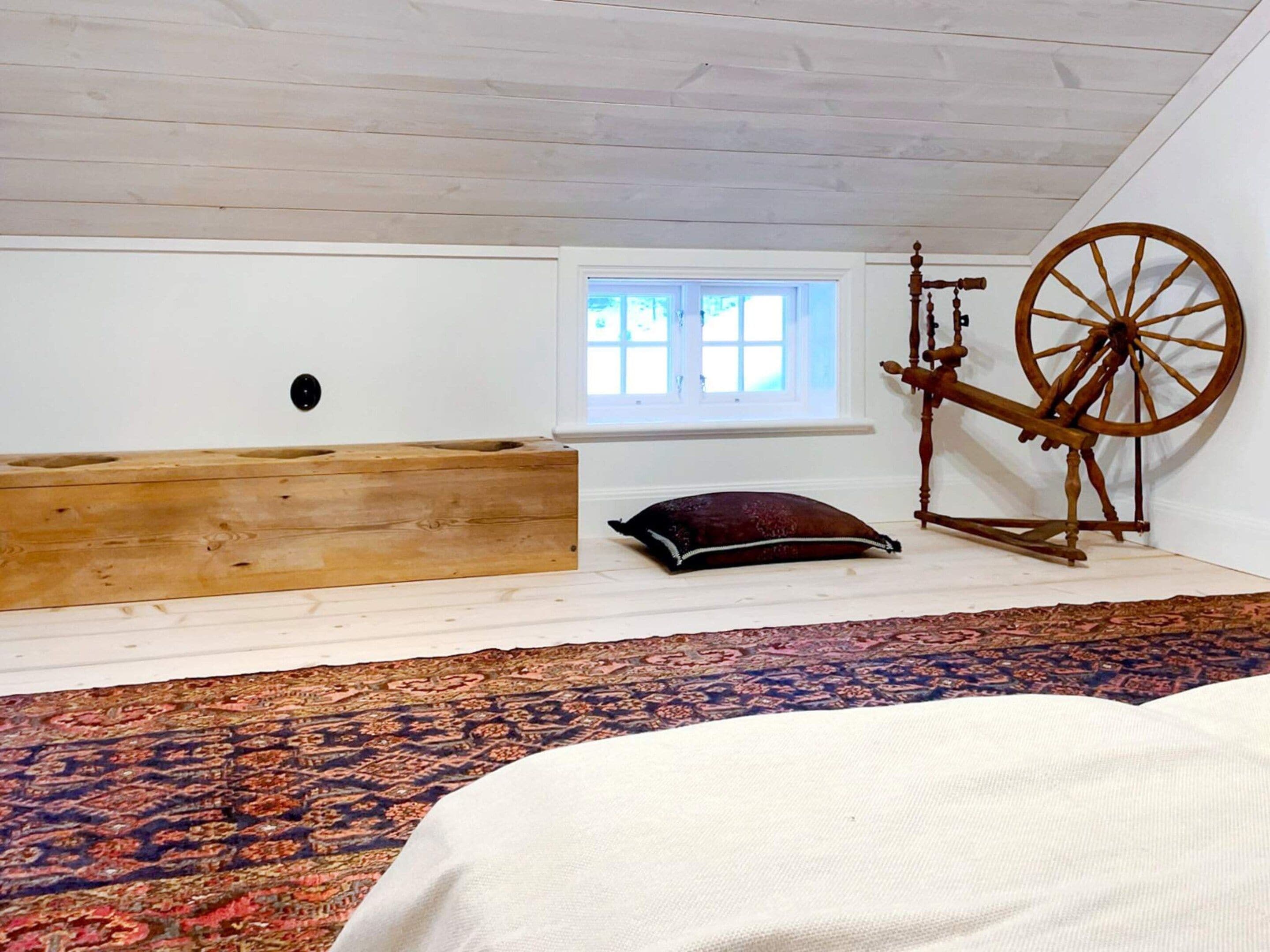 Een kamer met een schuine kap, een houten bankje, een spinnenwiel, een raam en een kamerbreed tapijt met dessin.