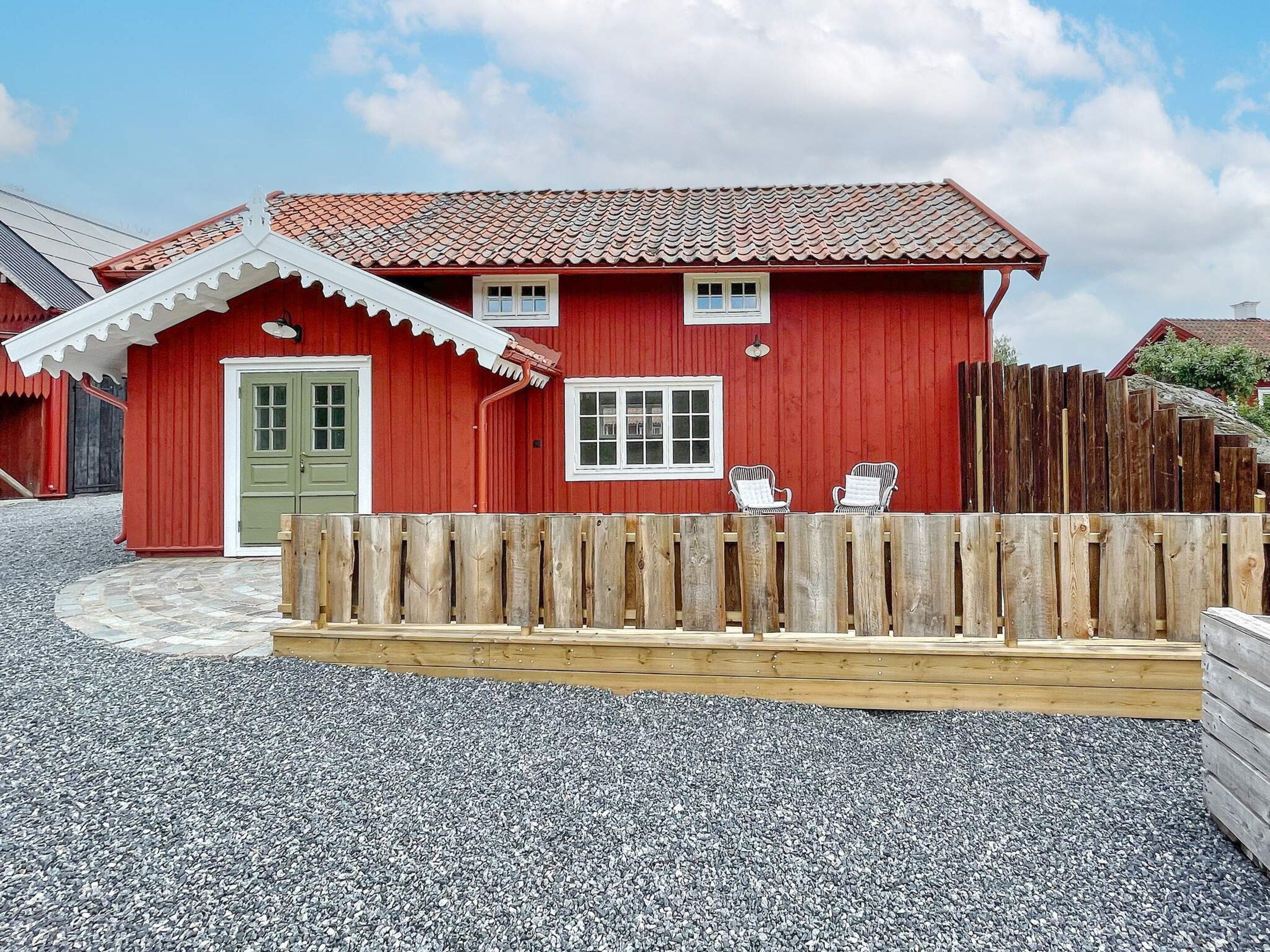 A red wooden house with a green door, two windows, and a wooden fence with two chairs.