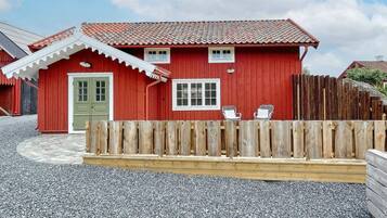 A red wooden house with a green door, two windows, and a wooden fence with two chairs.