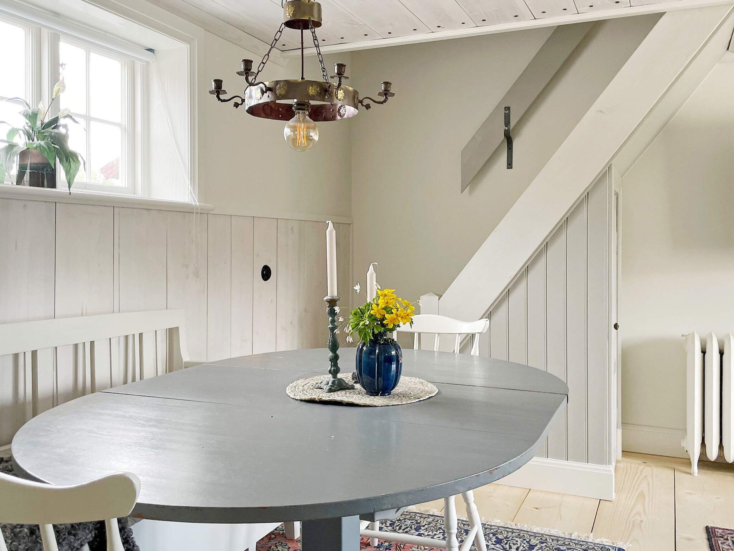 Un coin repas avec une table ronde, des chaises blanches, un lustre, un vase de fleurs et un escalier en arrière-plan.