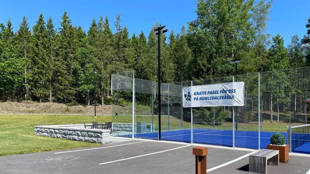 Un complexe sportif doté d’un terrain bleu, d’un panneau indiquant un service de navette gratuit et d’une zone délimitée par une clôture, avec des arbres en arrière-plan.