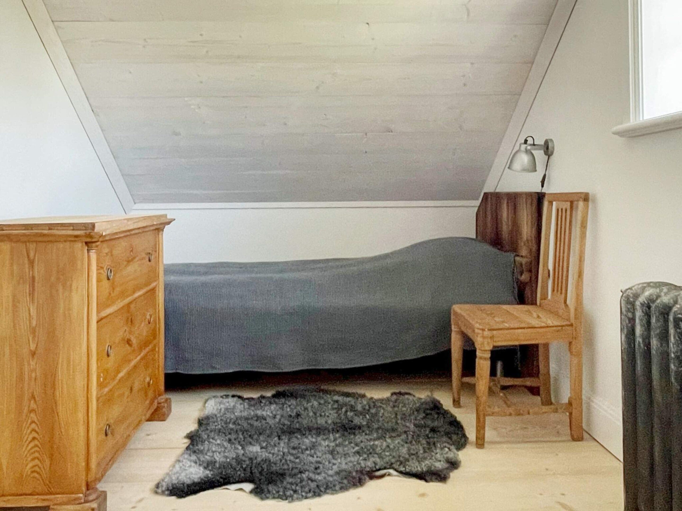Une petite chambre avec un lit simple, un plafond en pente, une commode en bois, une petite table et un tapis en fourrure sur le sol.