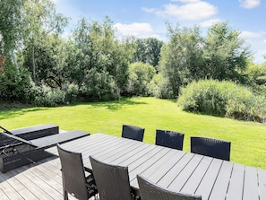 An outdoor patio with a wooden table and wicker chairs, surrounded by a grassy lawn and dense trees. - 5 Star Holiday Home in Vejby (Vejby)