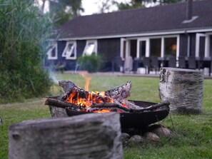 A campfire in a metal bowl with logs, surrounded by rocks, in a grassy outdoor area with a building in the background. - 5 Star Holiday Home in Vejby (Vejby)