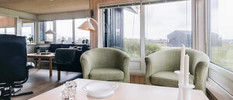Een kamer met een eettafel, twee fauteuils, een bank en een groot raam met uitzicht op de omgeving.