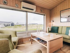 A room with a wooden interior, a large window with a view of the outdoors, a green sofa, a wooden coffee table, and a wall-mounted air conditioner. - 4 Person Holiday Home in Lokken (Lokken)