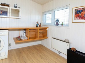 A compact kitchen with a washing machine, a wooden cabinet, a radiator, and a window with blinds.