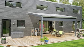 En moderne bygning med to etager, en overdækket terrasse, udendørs siddepladser og en velplejet græsplæne.