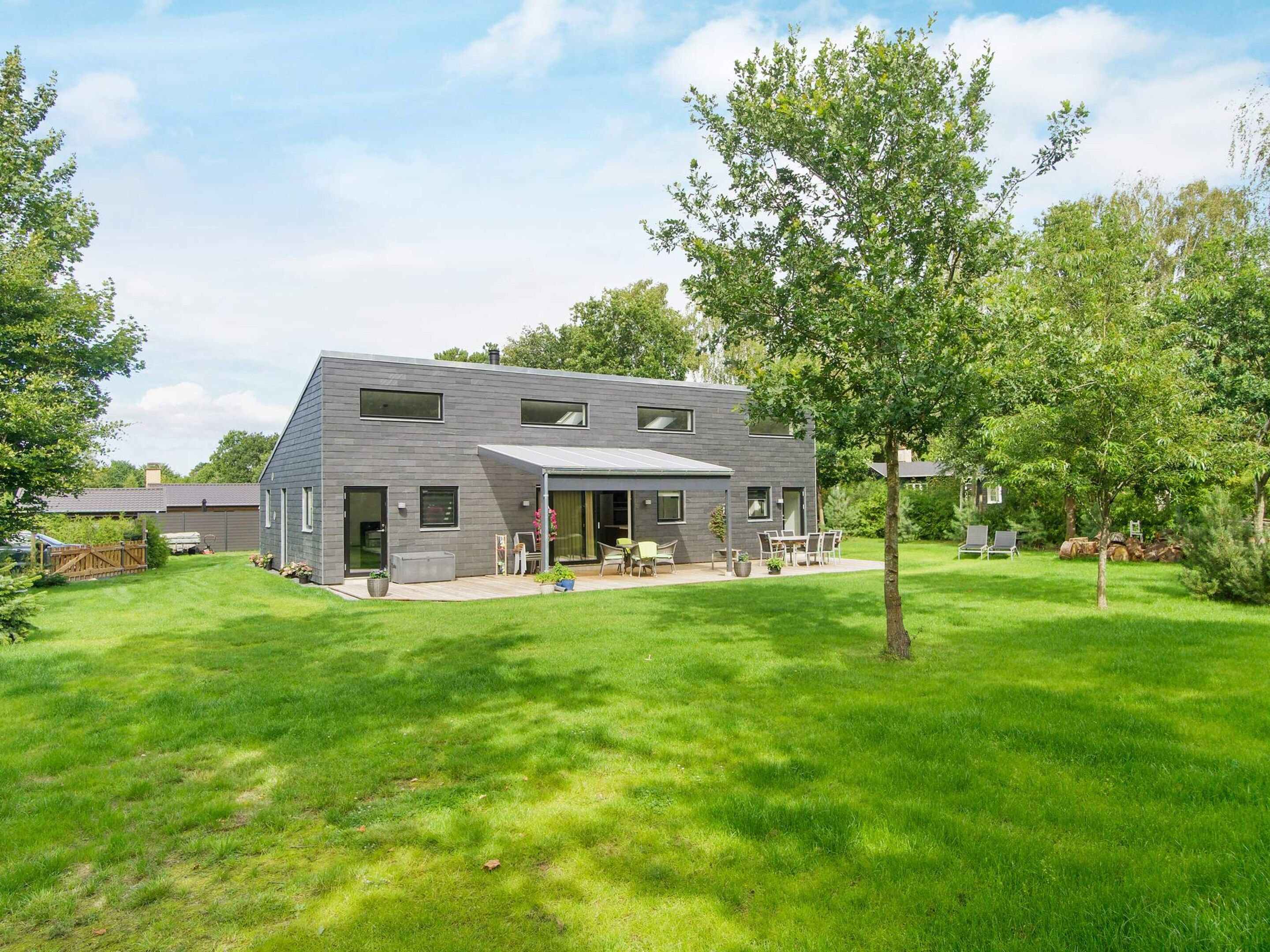 Et moderne hus i to etager med en overdækket terrasse, omgivet af en velholdt græsplæne og træer.