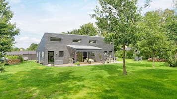 Et moderne hus i to etager med en overdækket terrasse, omgivet af en velholdt græsplæne og træer.