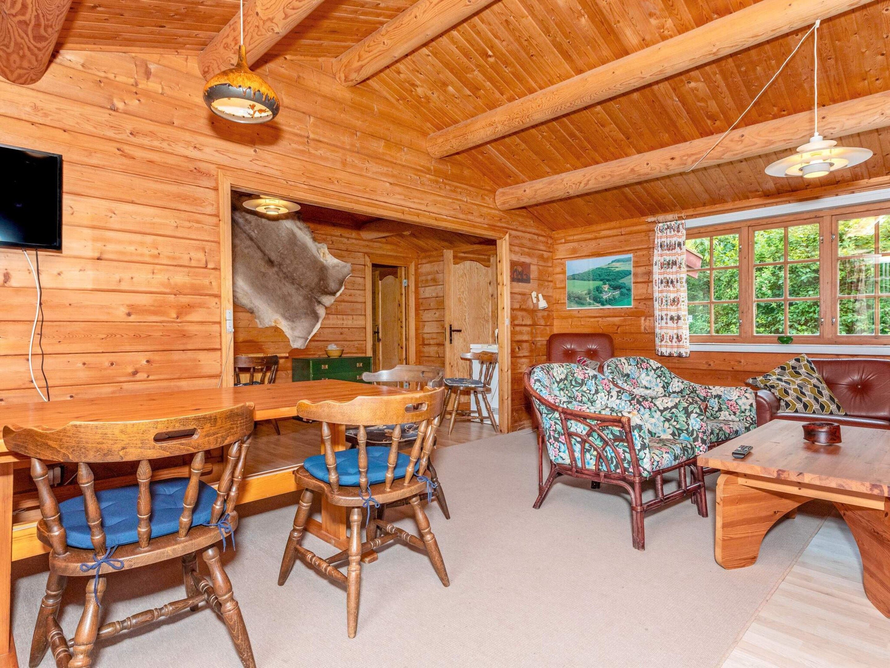 A wooden cabin interior with a dining table, chairs, a sofa, and a television mounted on the wall.