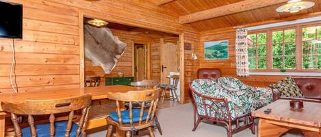 Interior de una cabaña de madera con una mesa de comedor, sillas, un sofá y un televisor montado en la pared.