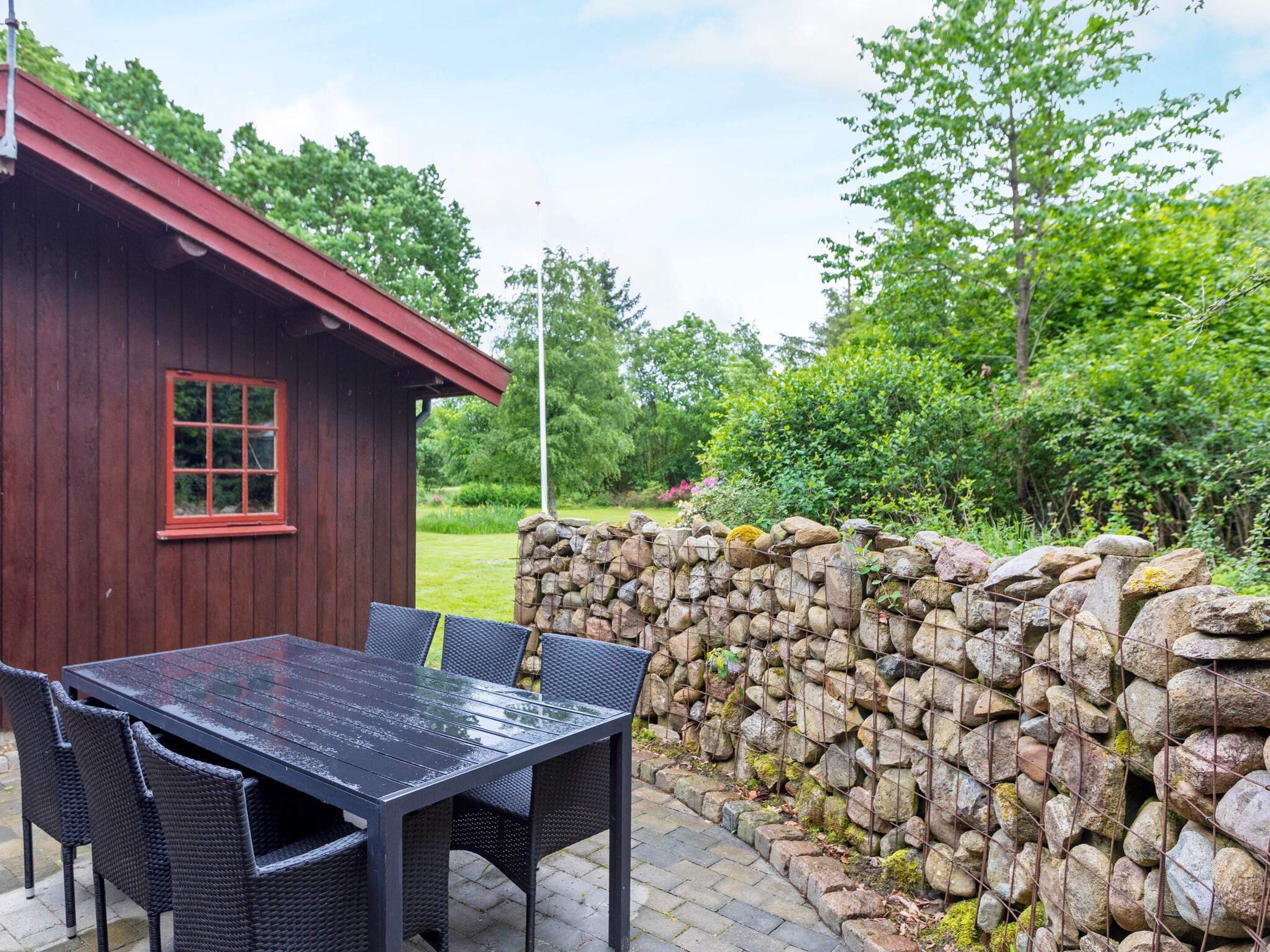 A wooden cabin with a red roof, a stone wall, and a table with chairs outside.