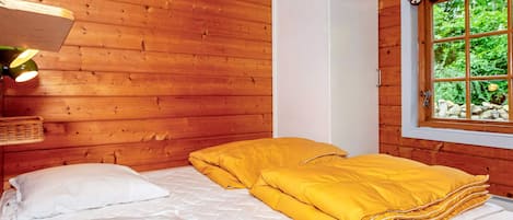 A wooden cabin room with a bed, a hanging light fixture, and a window with a view of greenery.