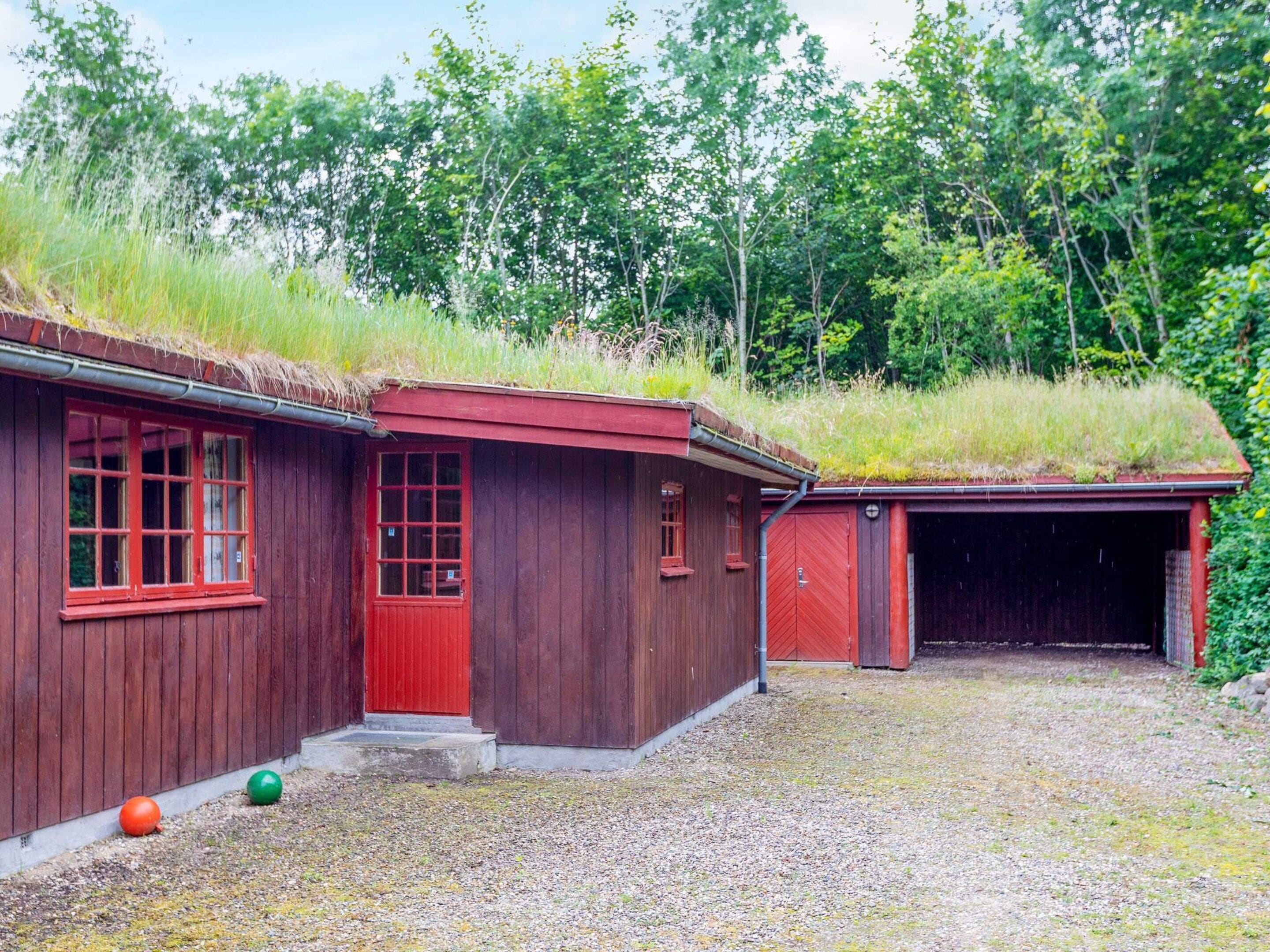 A wooden cabin with a red door and windows, a covered garage, and a grassy roof.
