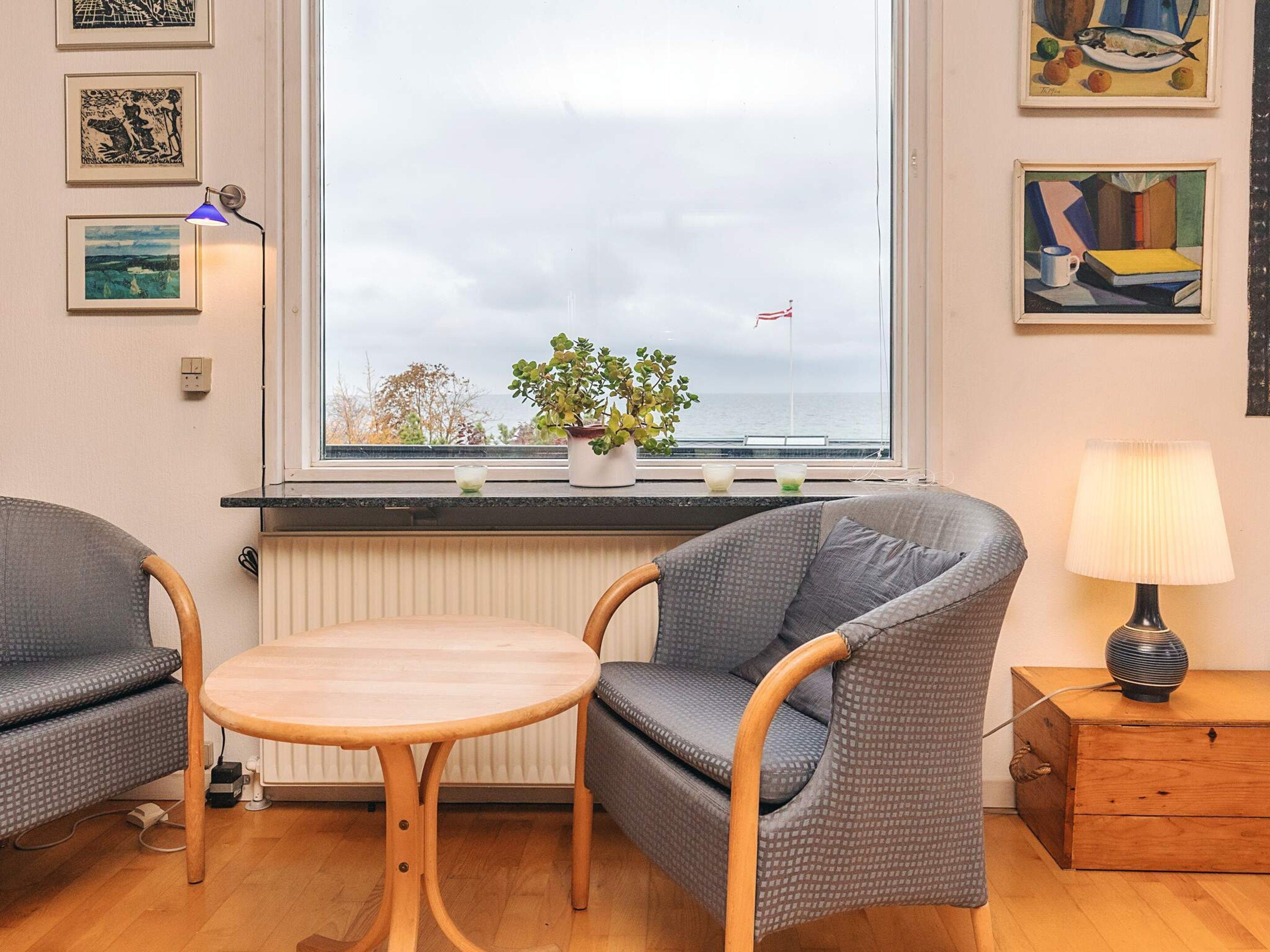 A cozy room with a large window, a round wooden table, two grey upholstered chairs, a small wooden nightstand with a lamp, and artwork on the walls.