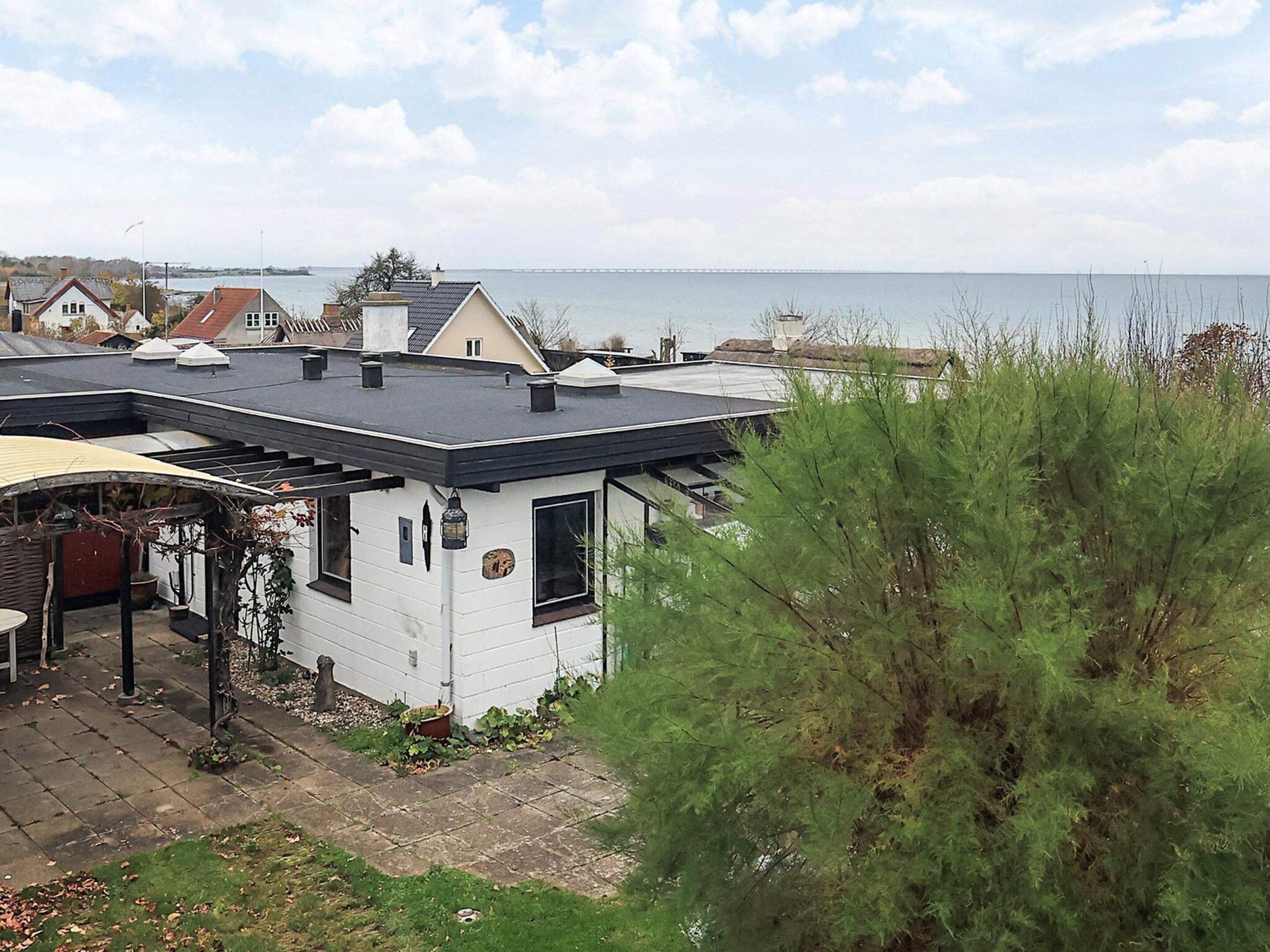 A white house with a black roof, a garden, and a view of the sea and a bridge in the background.
