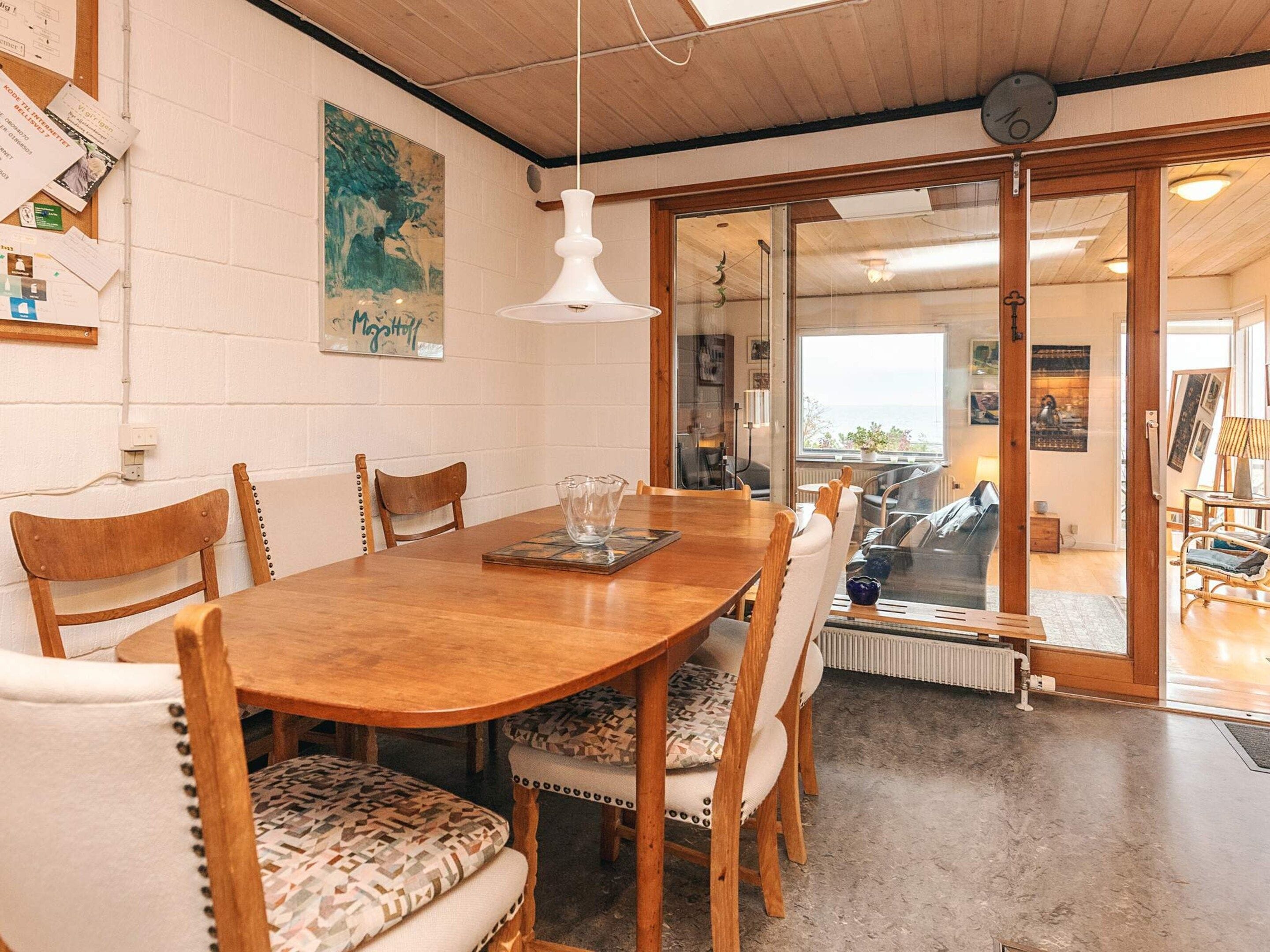 Ein Essbereich mit Holztisch und Stühlen, einer weißen Pendelleuchte und Blick durch eine Glastür in einen Wohnbereich mit Sofa und Lampe.
