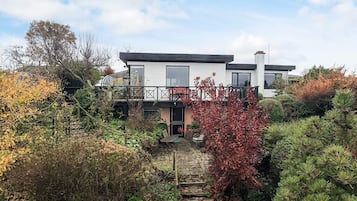 Ein modernes, zweistöckiges Haus mit Balkon, umgeben von einem Garten mit verschiedenen Pflanzen und Bäumen.