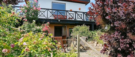 Una casa moderna de dos pisos con balcón, rodeada de jardines exuberantes y plantas en flor.