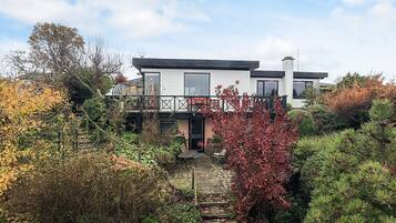 Ein modernes, zweistöckiges Haus mit Balkon, umgeben von einem Garten mit verschiedenen Pflanzen und einem Kiesweg, der zum Eingang führt.