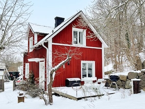 A red wooden house with a chimney, a porch with chairs, and a snow-covered yard.