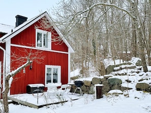 A red house with a chimney, a window, and a door. There is a snow-covered ground and trees in the background.